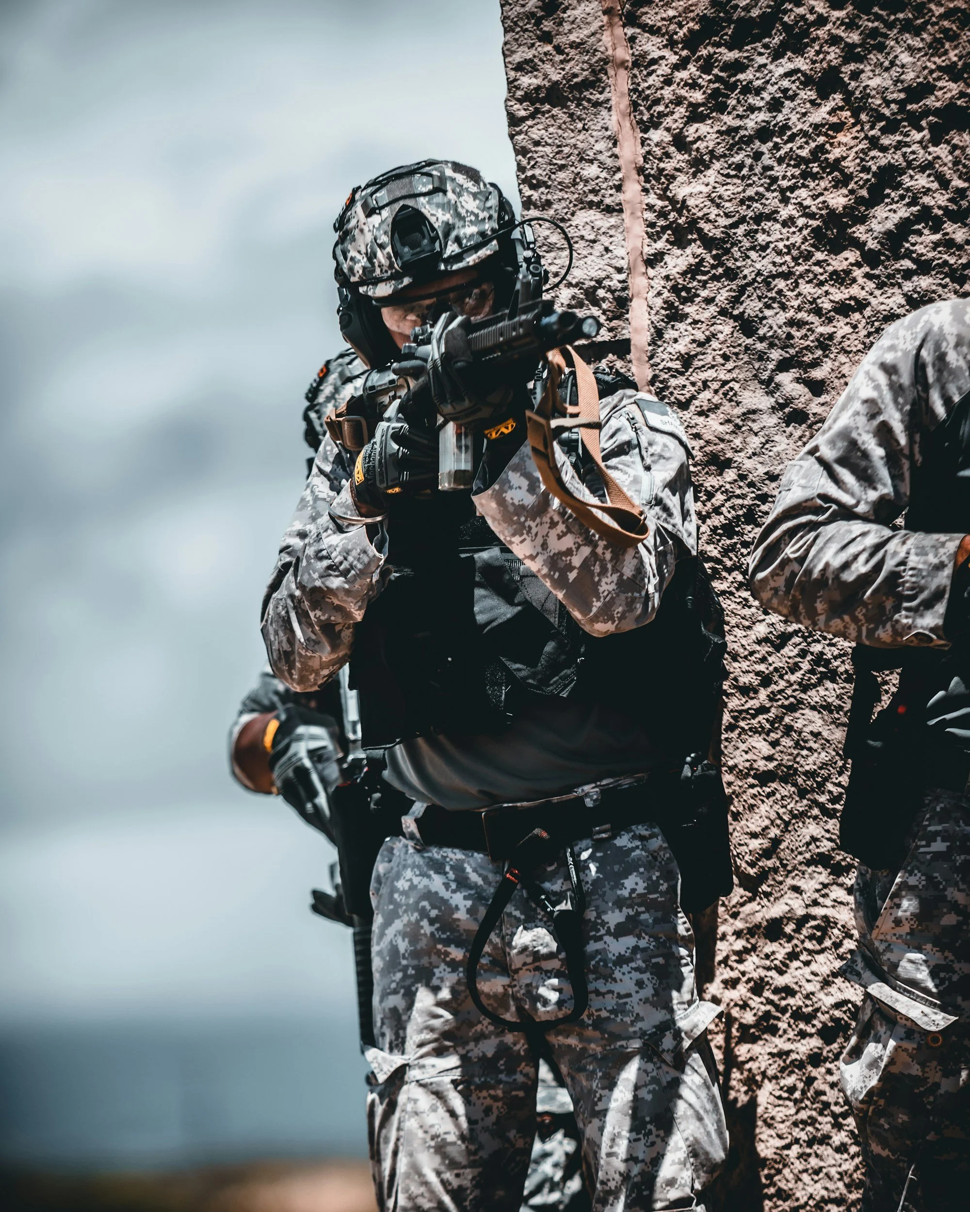Military soldier in camouflage uniform aiming a firearm during training or combat, with cloudy sky in background.