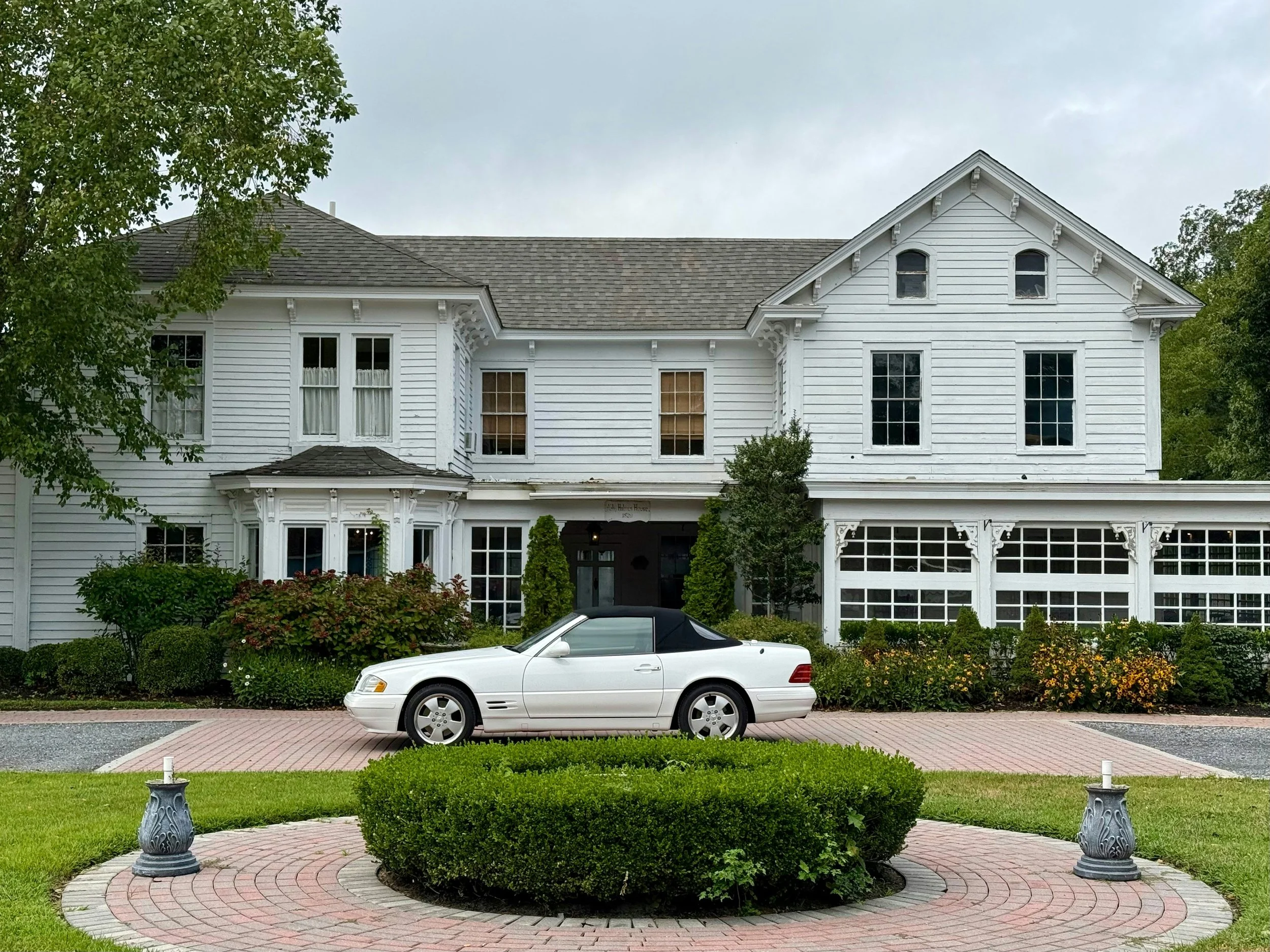 Front view of a large white house with a black convertible car parked in driveway, surrounded by green bushes and trees, overcast sky.