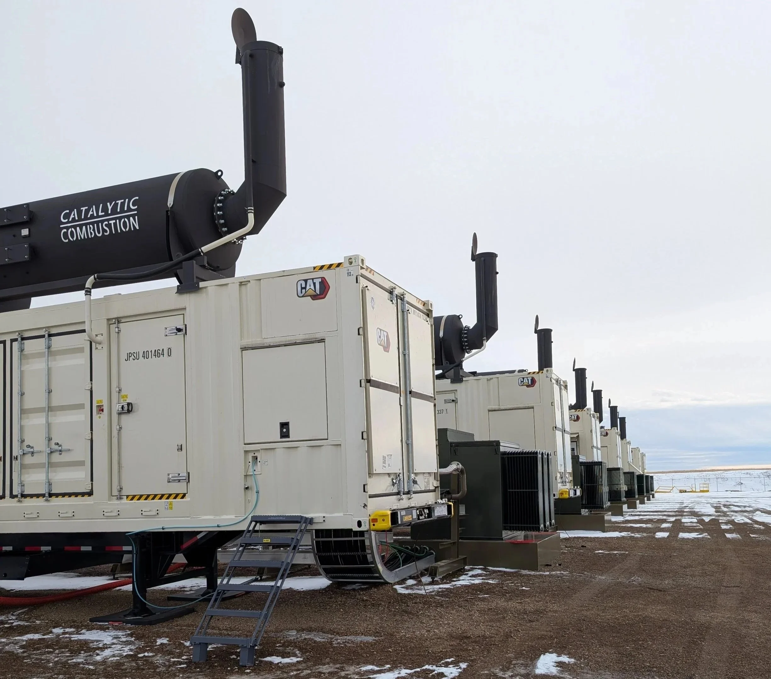 A row of large industrial generator units outdoors with exhaust stacks and equipment, some snow on the ground, cloudy sky.