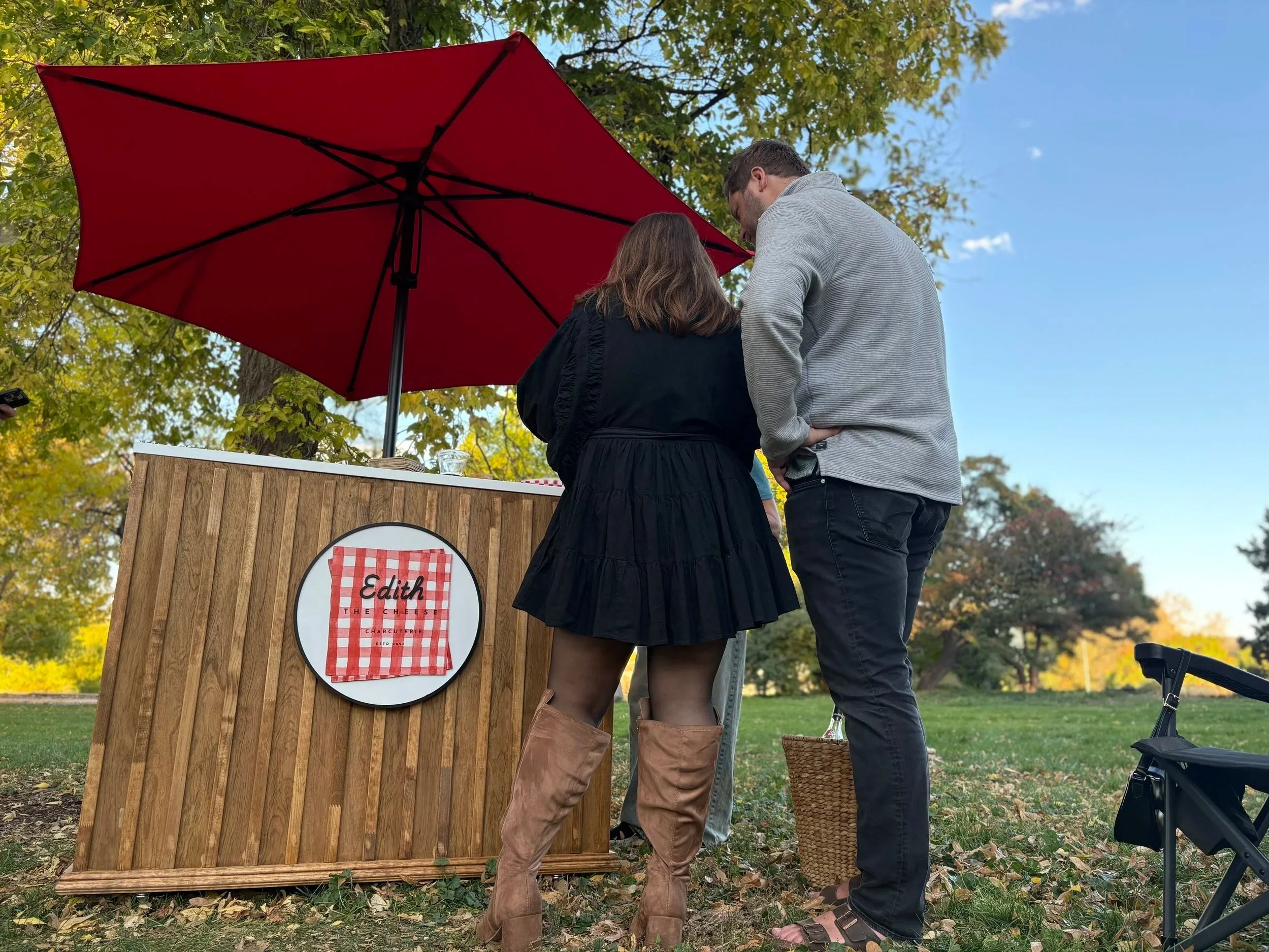 A small outdoor cheese and wine stand with a red umbrella in a park, featuring cheese trays, a bottle of wine, and a sign that reads 'Edith The Cheese' with a red and white checkered cloth design.