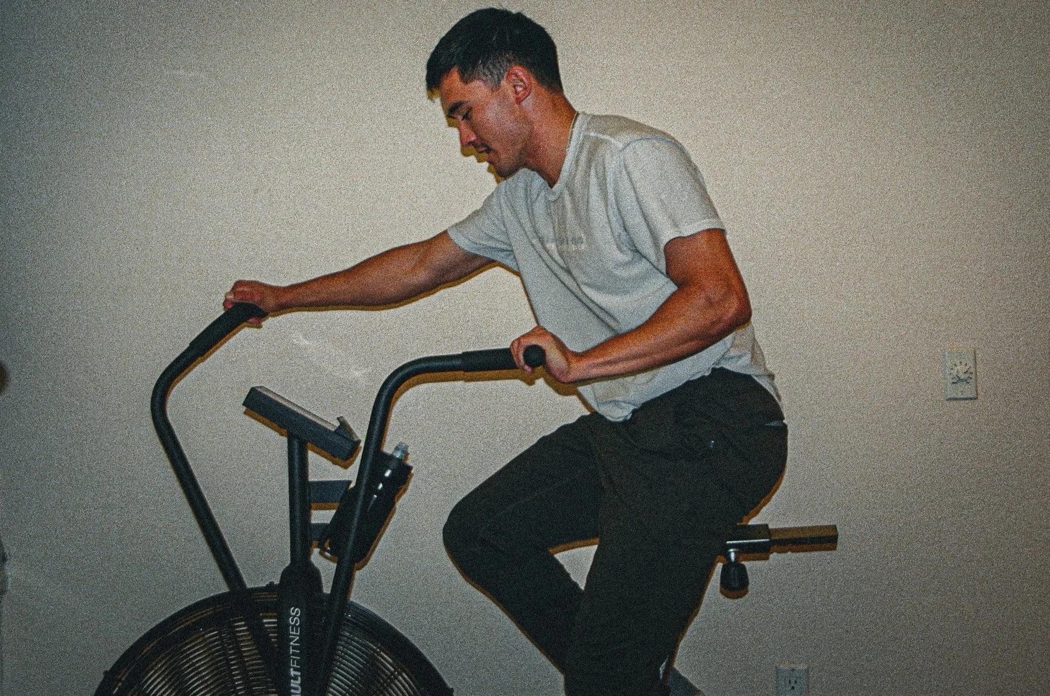 A young man is riding an exercise bike indoors, wearing a gray T-shirt and black pants, with a plain wall in the background.