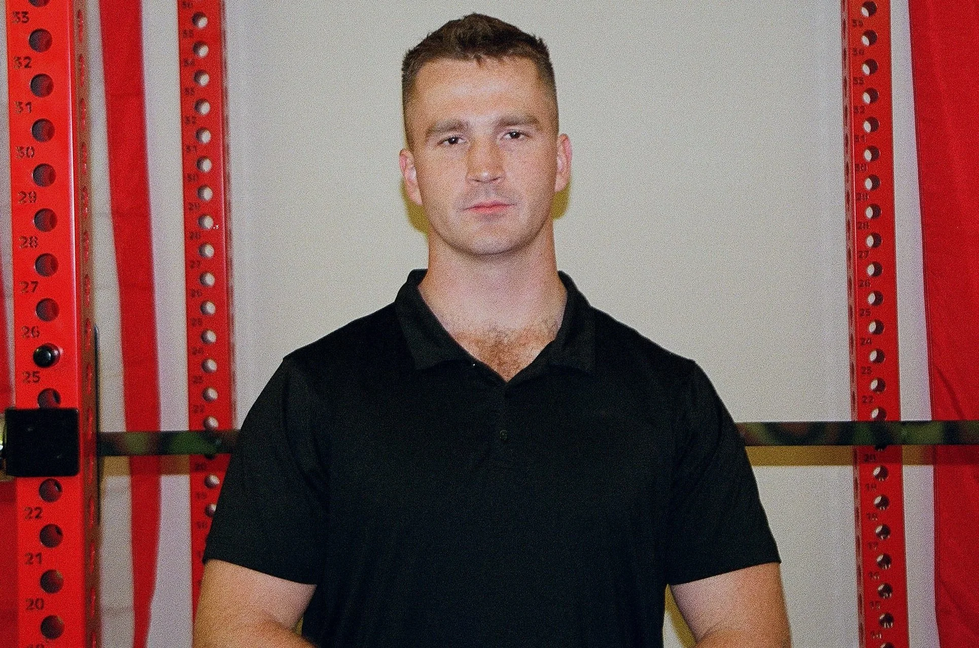 A young man with short brown hair and fair skin standing against a white background with red and white vertical stripes on either side. He is wearing a black polo shirt and has a serious expression.