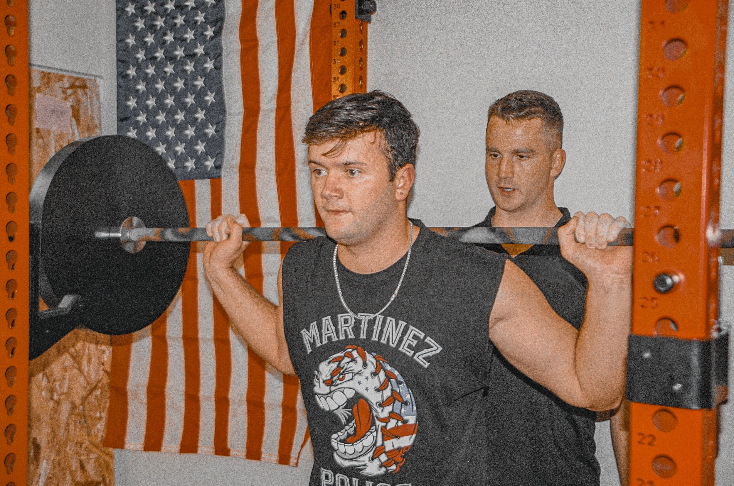 Two men lifting a barbell in a gym with an American flag on the wall.
