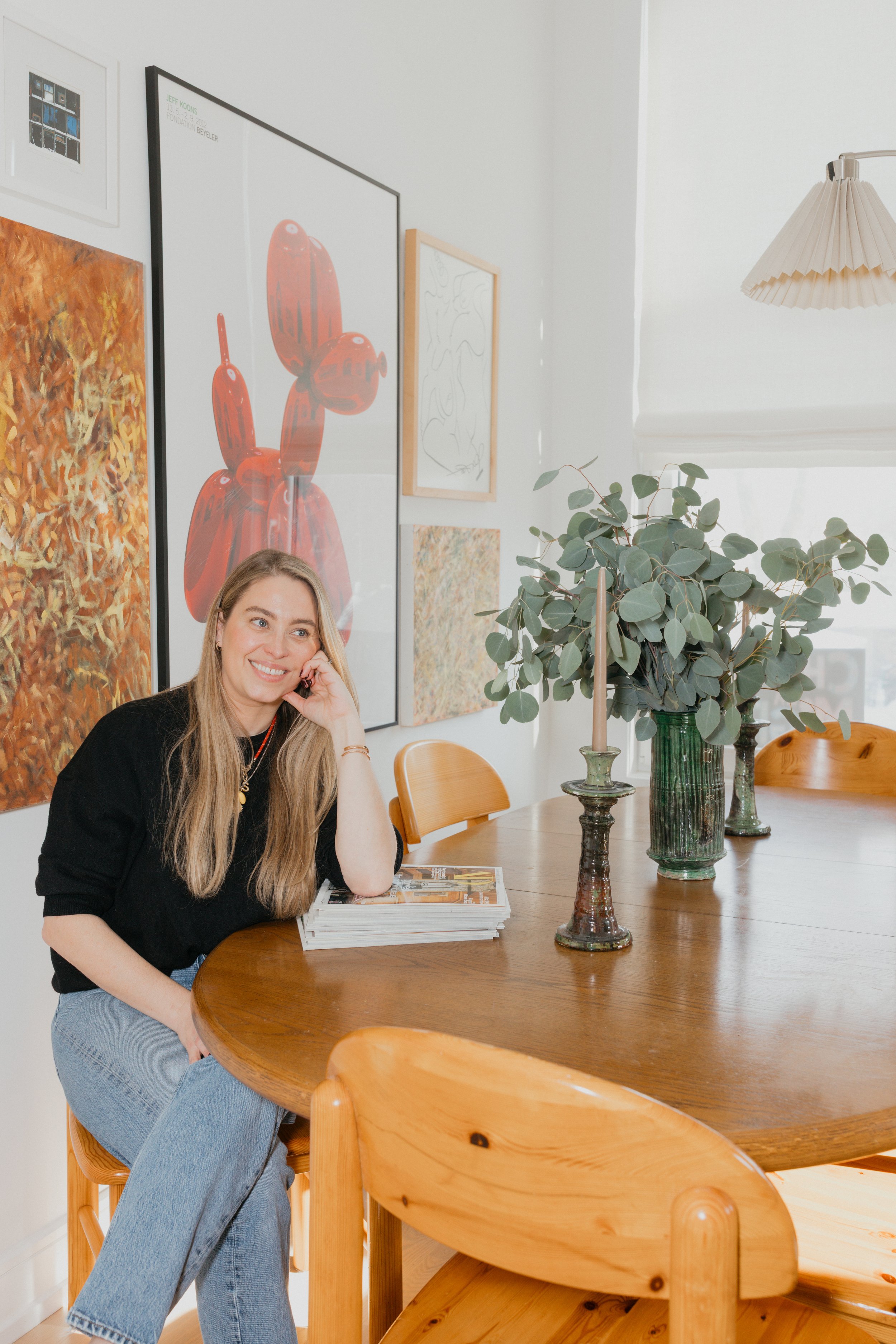 Founder Daniella Mayer wearing a black top and jeans, sitting at a wooden dining table, smiling, with art prints on the wall behind her and green foliage in a vase on the table.