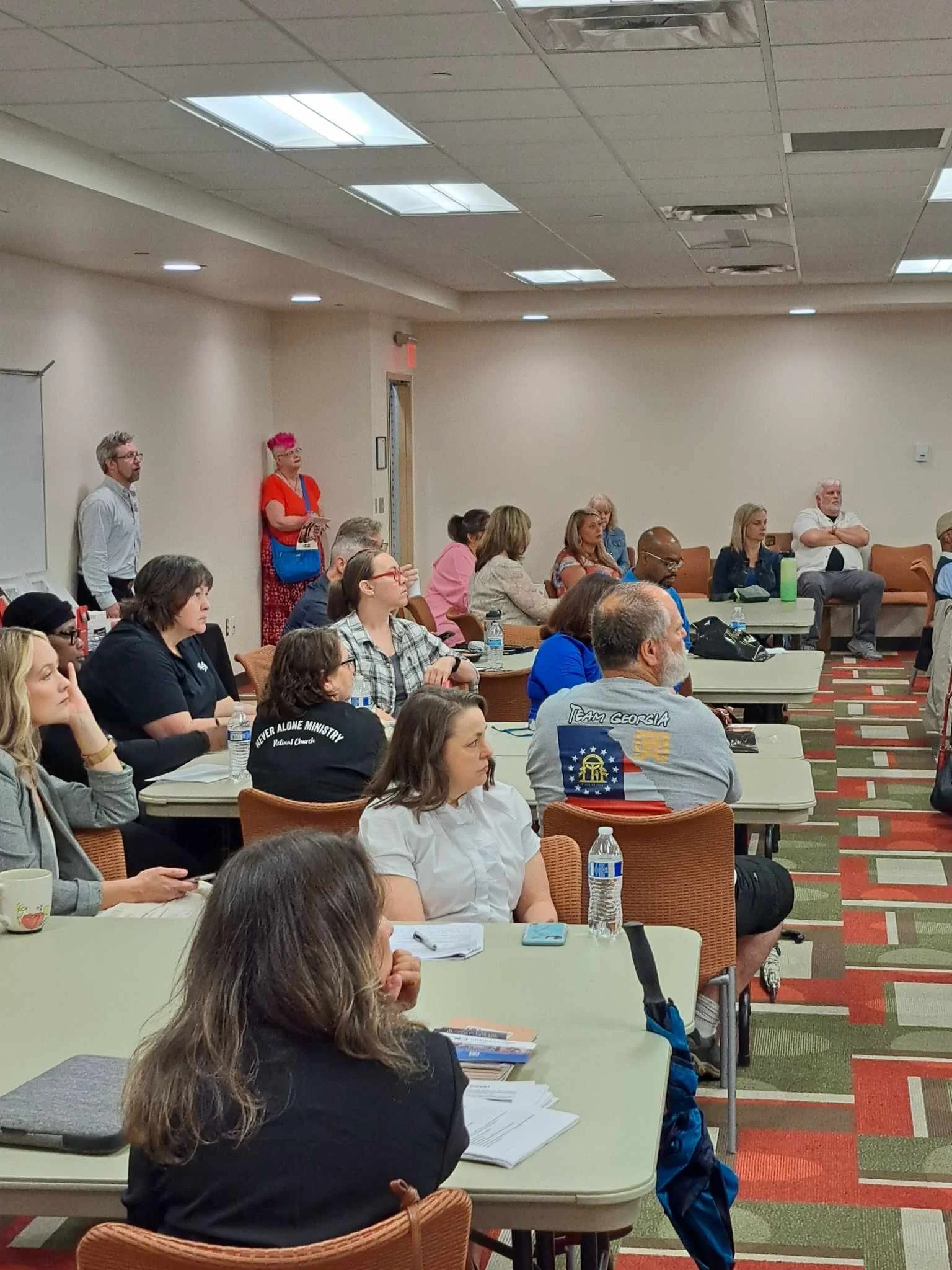 People seated at desks in a conference room, listening attentively. A woman stands near the wall, wearing a bright orange shirt and pink hair.