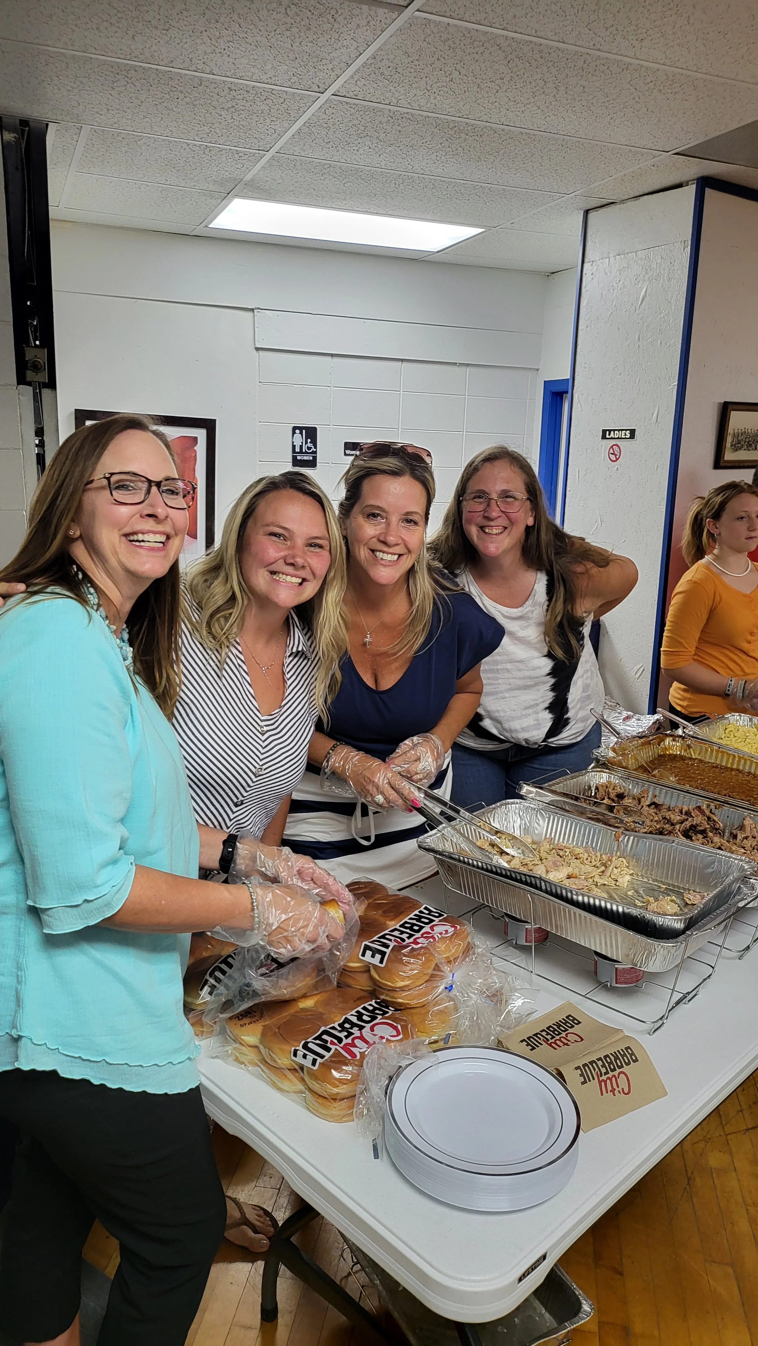 Four women smiling and serving food at a buffet table in a community center or hall, with trays of barbecue, buns, and plates on the table.