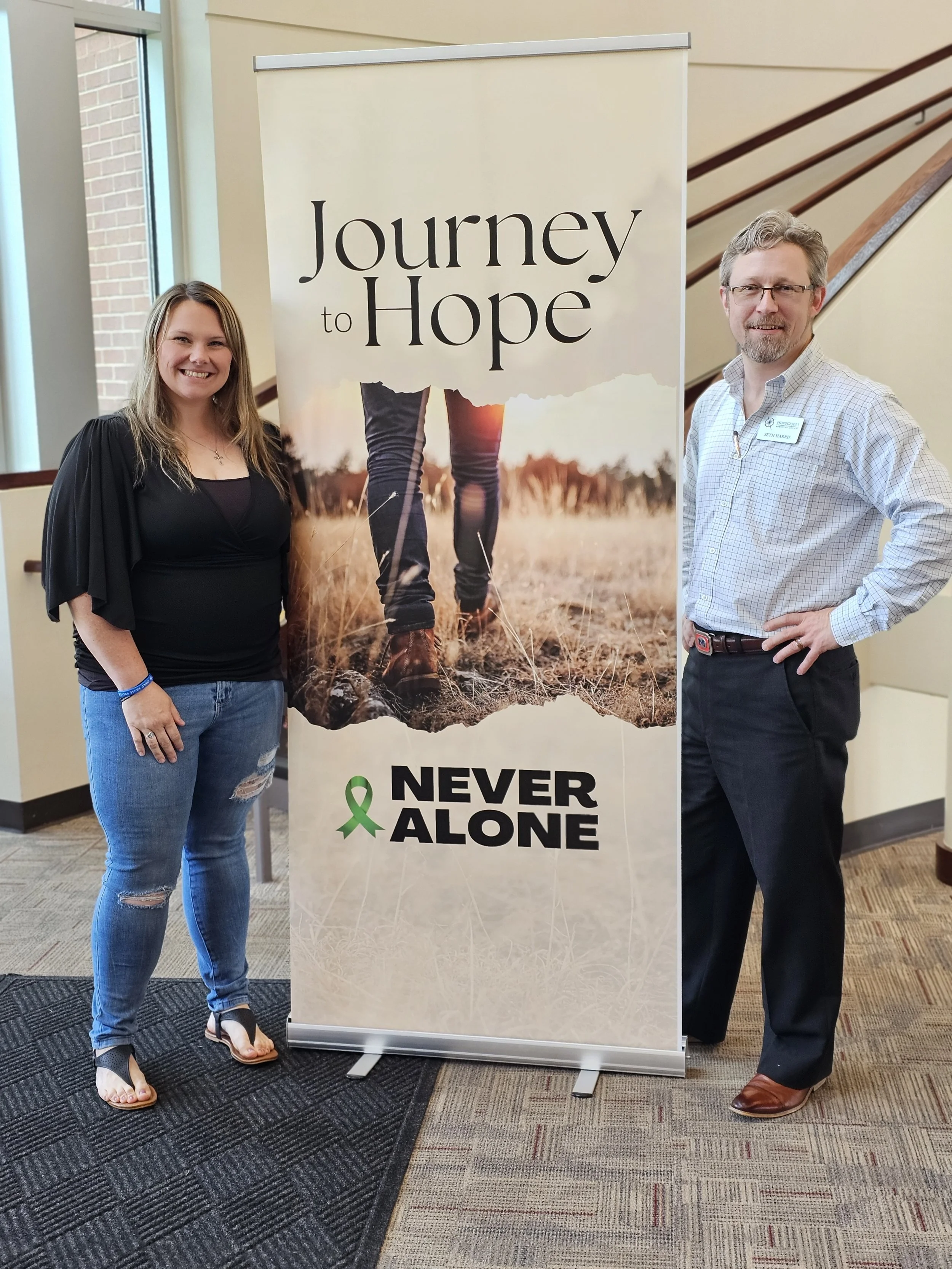 Two people standing next to a banner that reads 'Journey to Hope' and 'Never Alone', with an image of someone walking in a field.