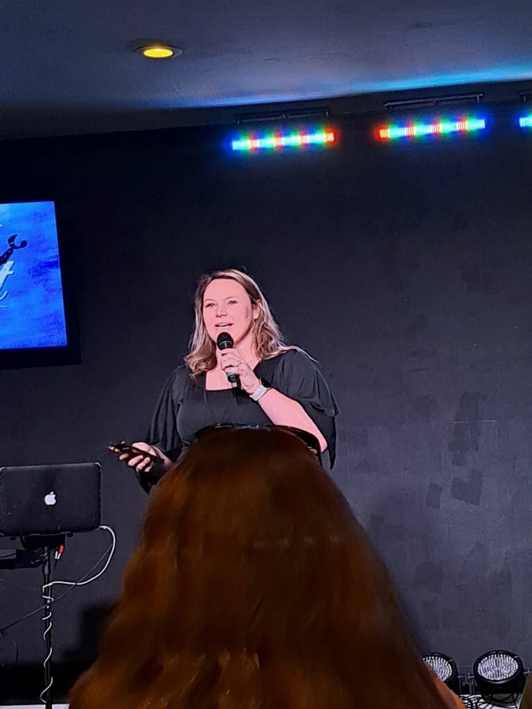 A woman is speaking on a stage with a microphone in her hand, standing in front of a black wall with colorful lights above her. There is a laptop on a stand and a screen to her left showing a blue graphic, and an audience member's head with brown hair is visible in the foreground.
