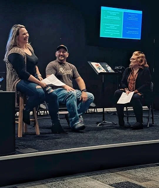 Three people participating in a panel discussion or workshop in a dark room with a large screen displaying presentation slides behind them. The woman on the left is standing and speaking, the man in the middle is sitting and smiling, and the woman on the right is sitting holding papers.