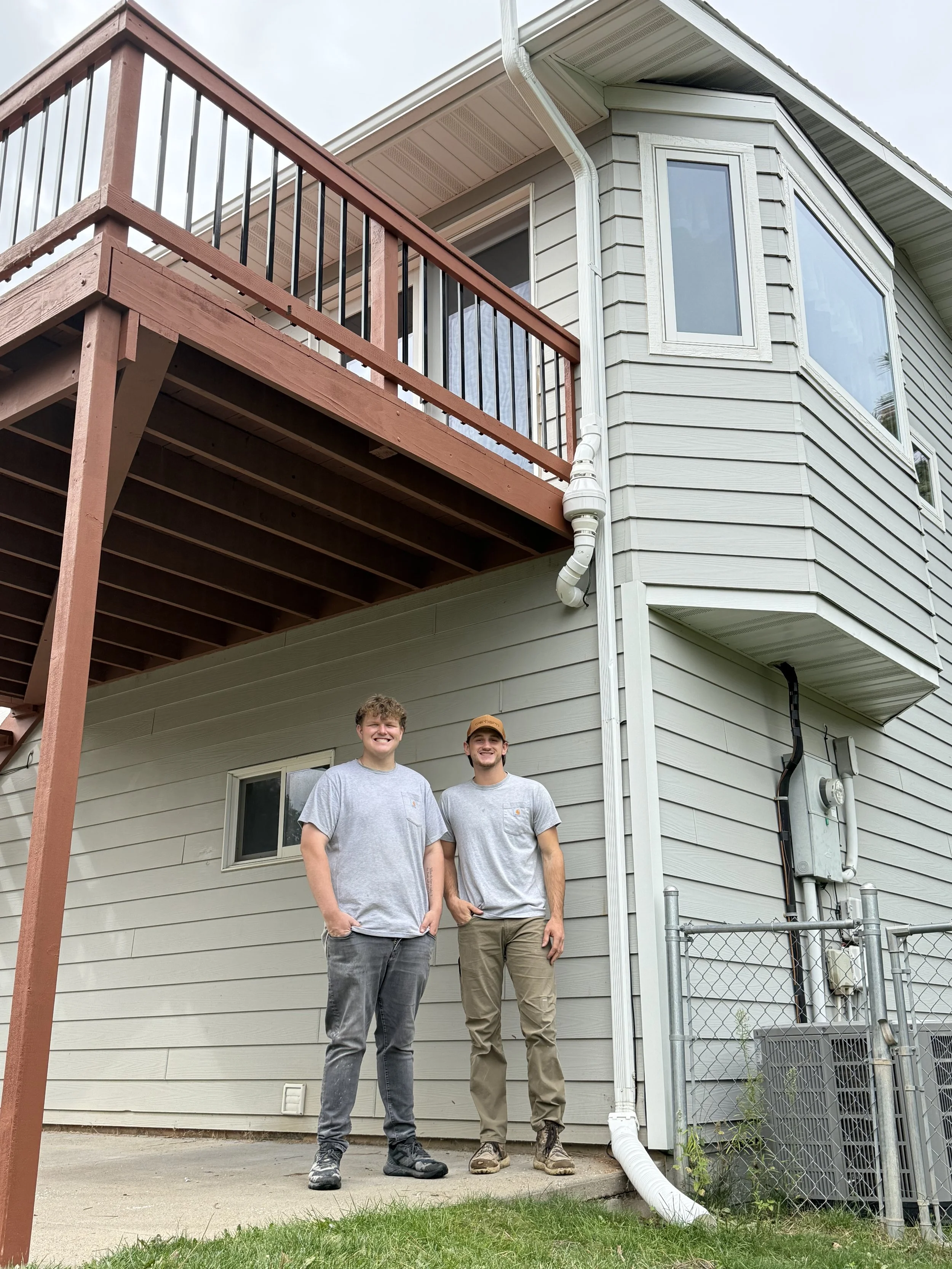 Two young men standing on a concrete patio in front of a light-colored house with a second-story balcony and siding, smiling at the camera.