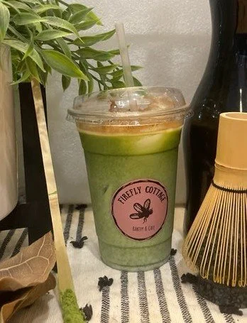 A green iced drink in a clear plastic cup with a straw, sitting on a black and white striped cloth, with a pink label reading 'Firefly Cottage' and surrounded by plants, bottles, and decorative items.