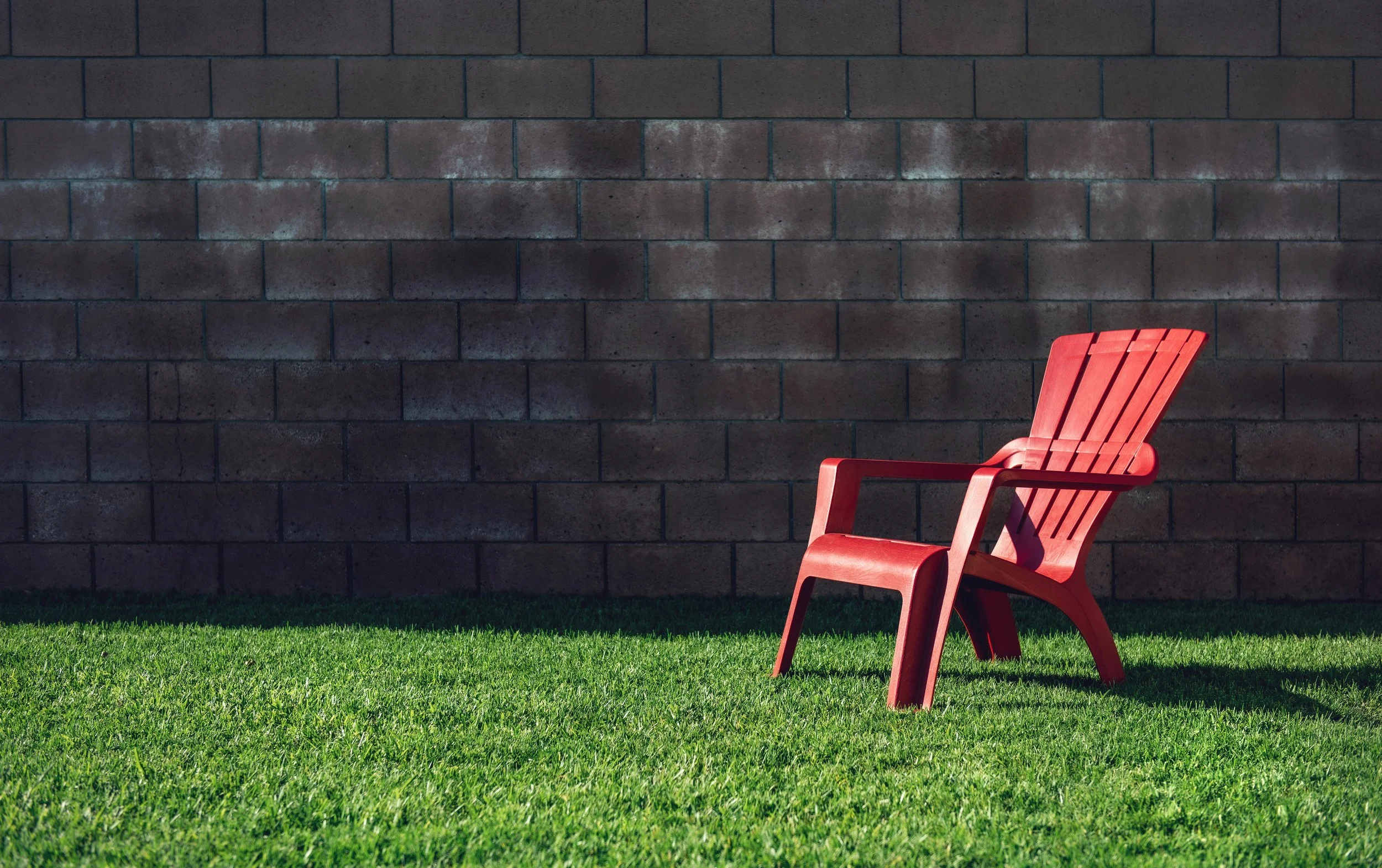 A red plastic outdoor chair on a grassy lawn with a brick wall background.