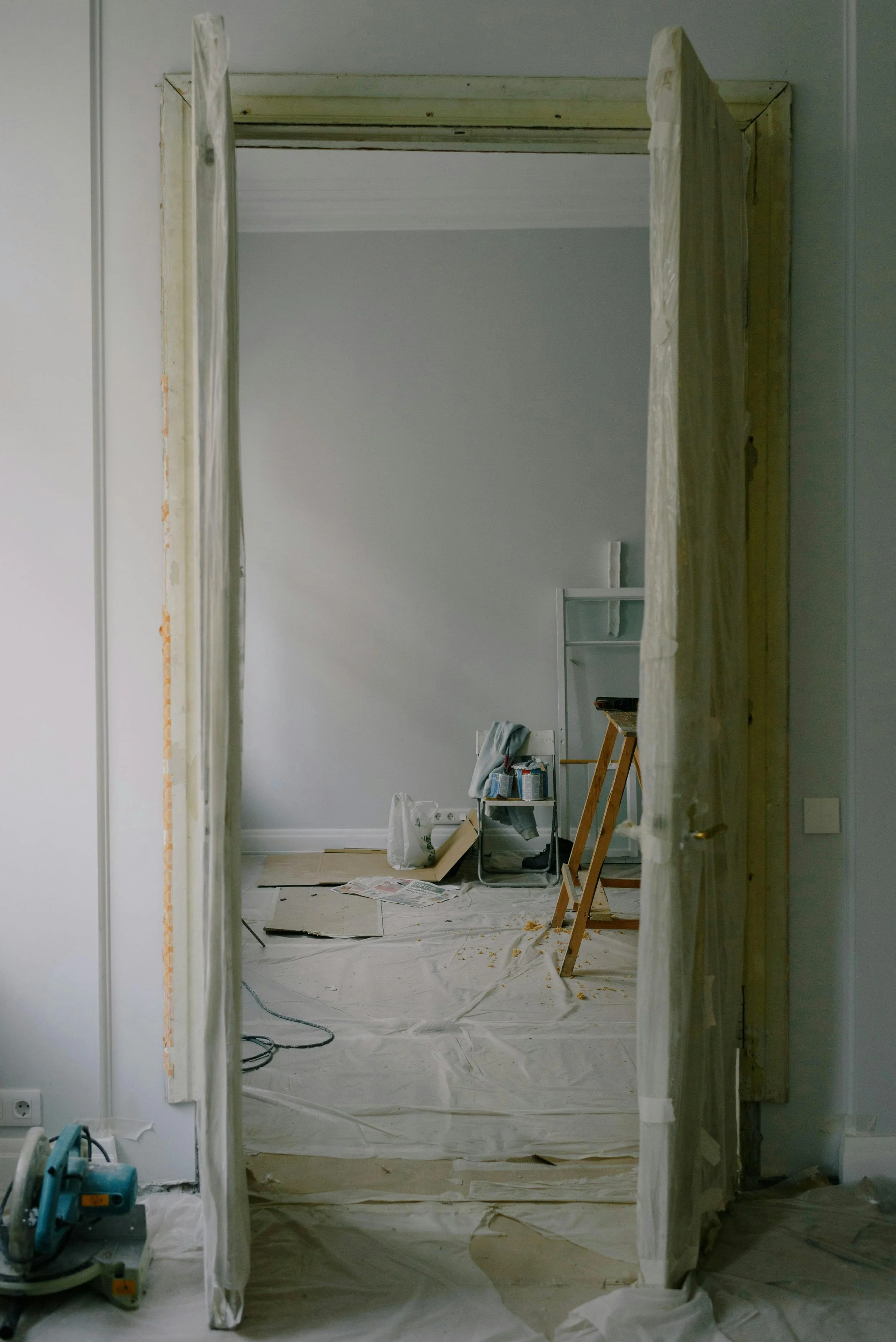 Interior of a room under renovation, viewed through an open doorway with curtains. The room has a light-colored wall, construction tools, a small table with painting supplies, and the floor covered with protective plastic sheeting.