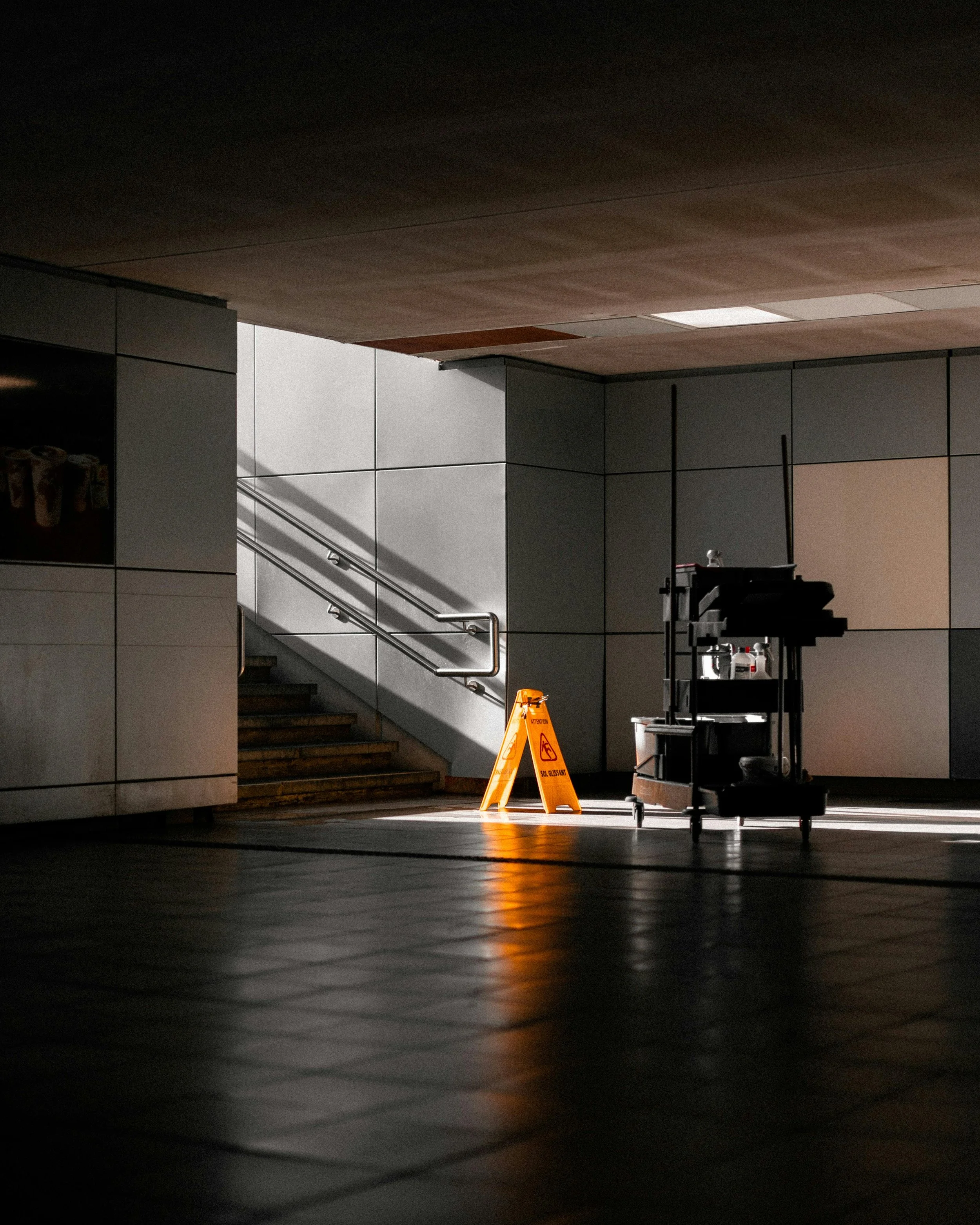 Empty indoor space with a caution sign, cleaning trolley, and stairs lit by sunlight.