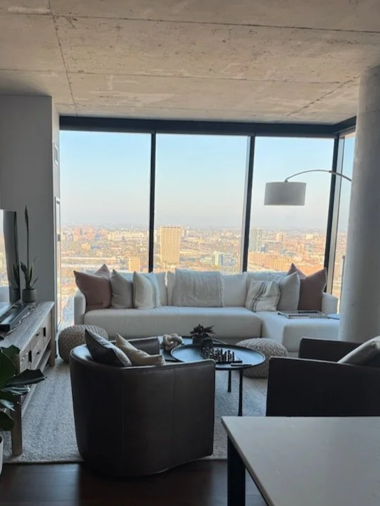Modern living room with large windows showing a cityscape, a white sofa with pillows, a black armchair, a round coffee table with decorative items, and a floor lamp.