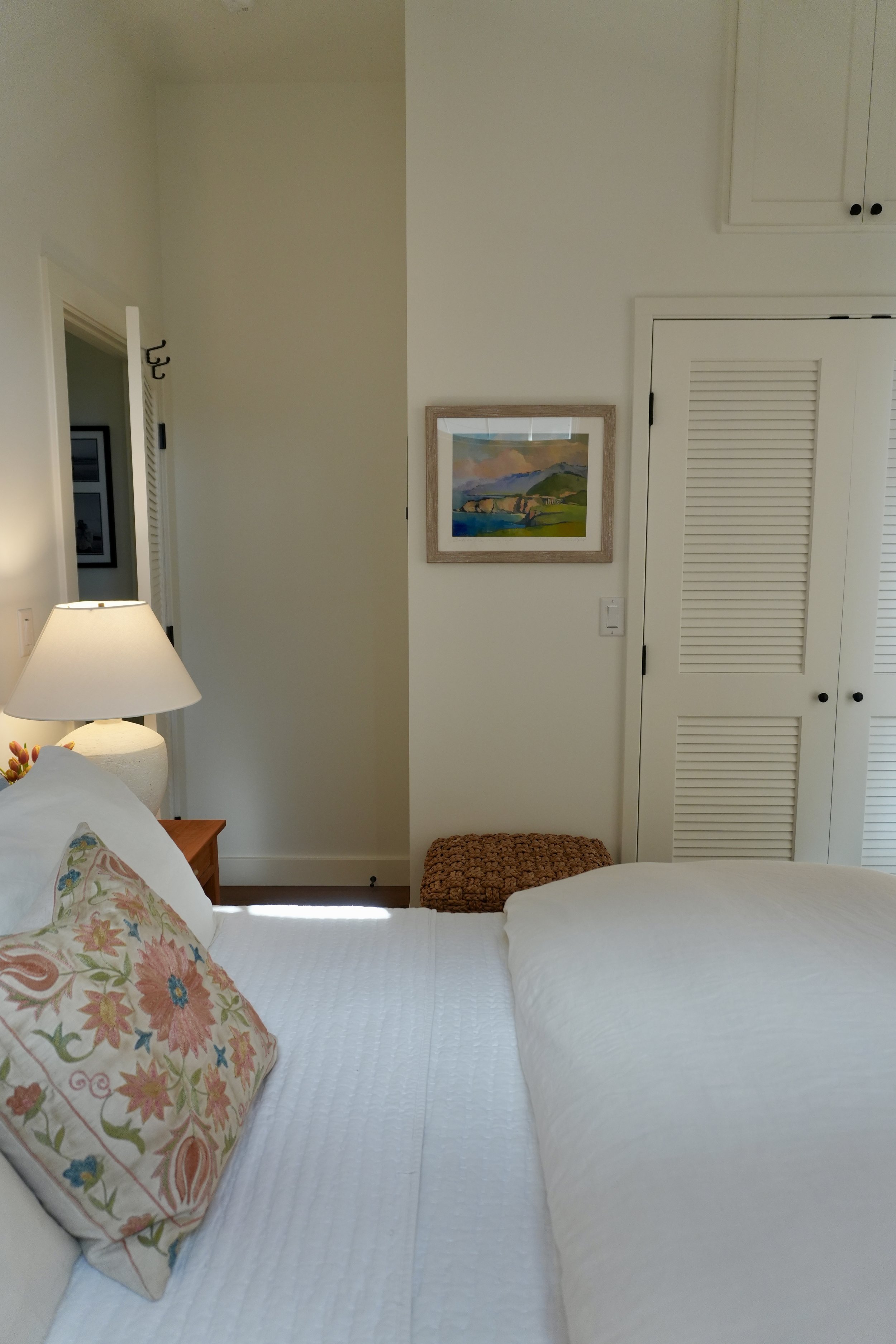 A bedroom with a partially visible bed, floral pillow, bedside lamp, framed landscape painting, and closet doors.