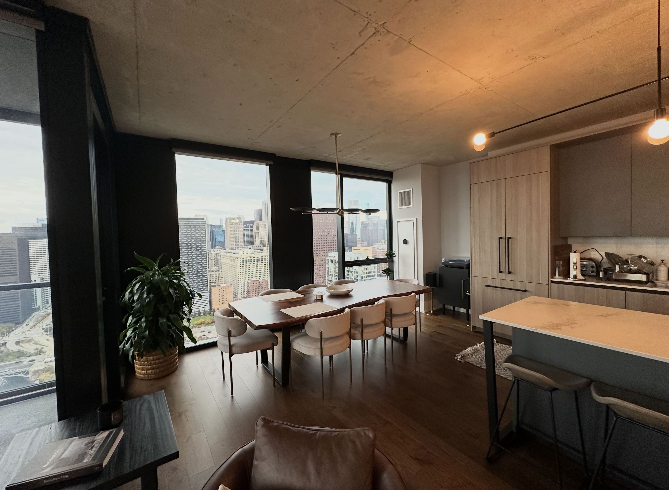 Modern high-rise apartment dining area with large windows, a wooden dining table, beige chairs, potted plant, and cityscape view.