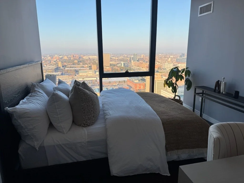 Bedroom with large window overlooking city skyline, bed with white and patterned pillows, minimal decor, and a small plant.