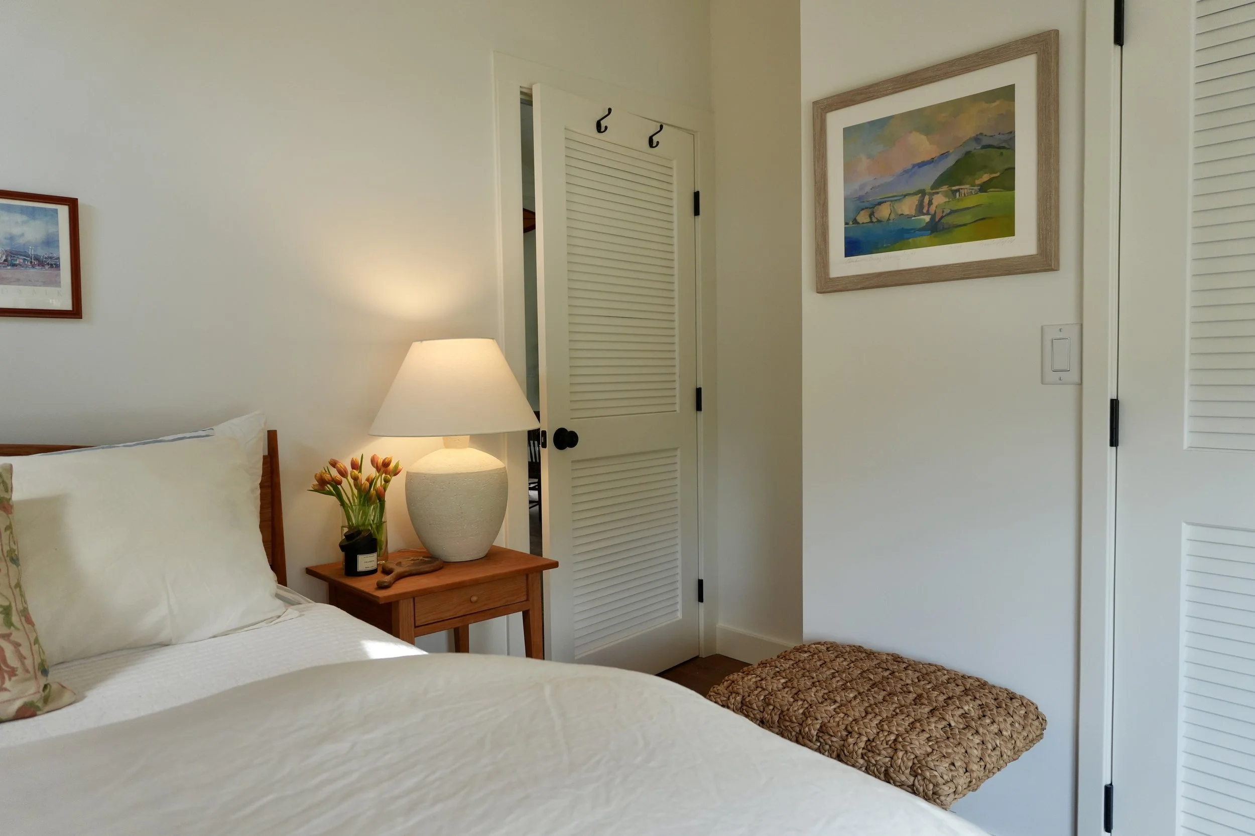 Part of a bedroom with a bed, pillow, nightstand with a lamp, flowers, and picture frames on the wall.