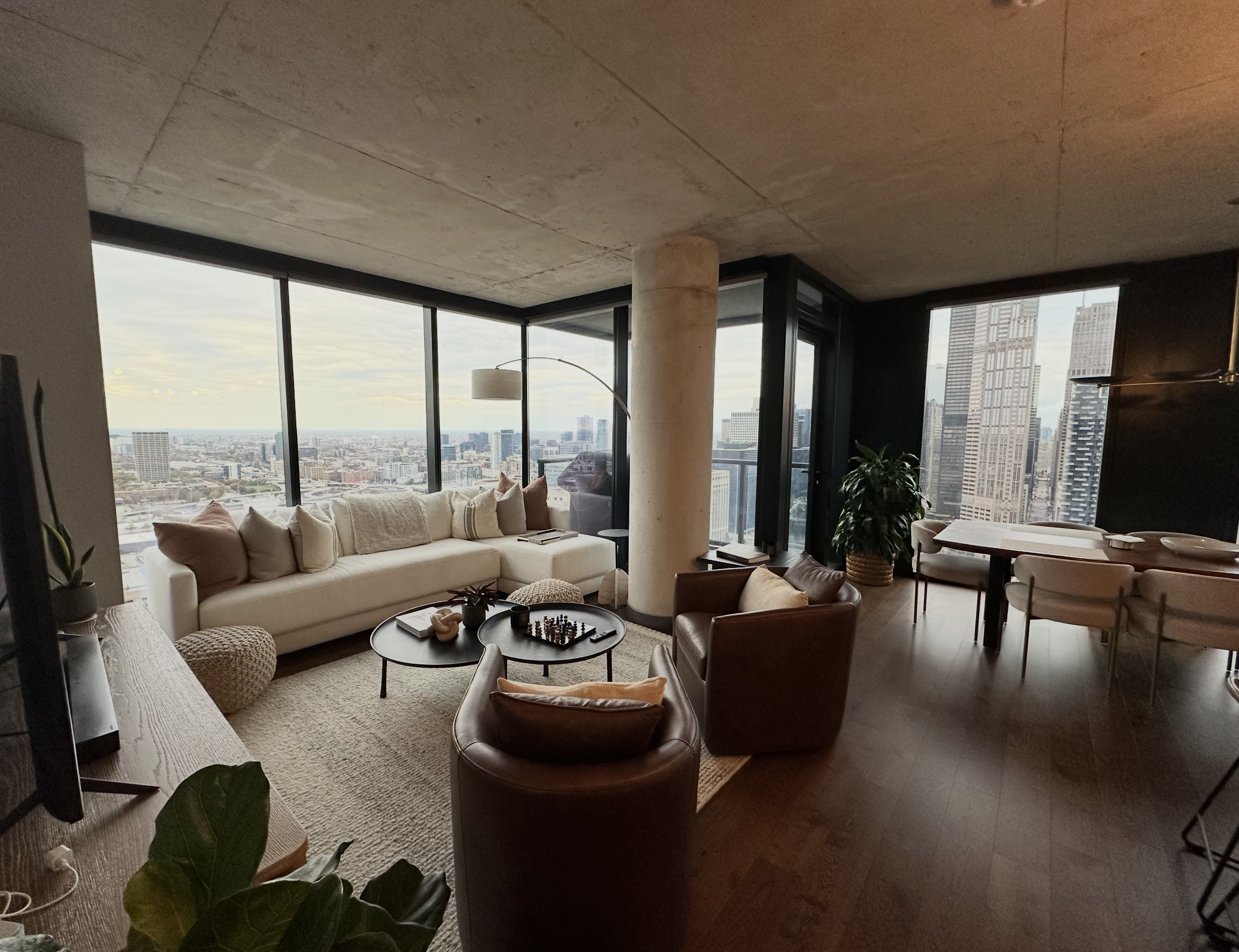 A modern high-rise apartment living room with large windows showing a city skyline, a beige sofa with cushions, a round coffee table with books and decorative items, two armchairs, a dining table with chairs, and indoor plants.