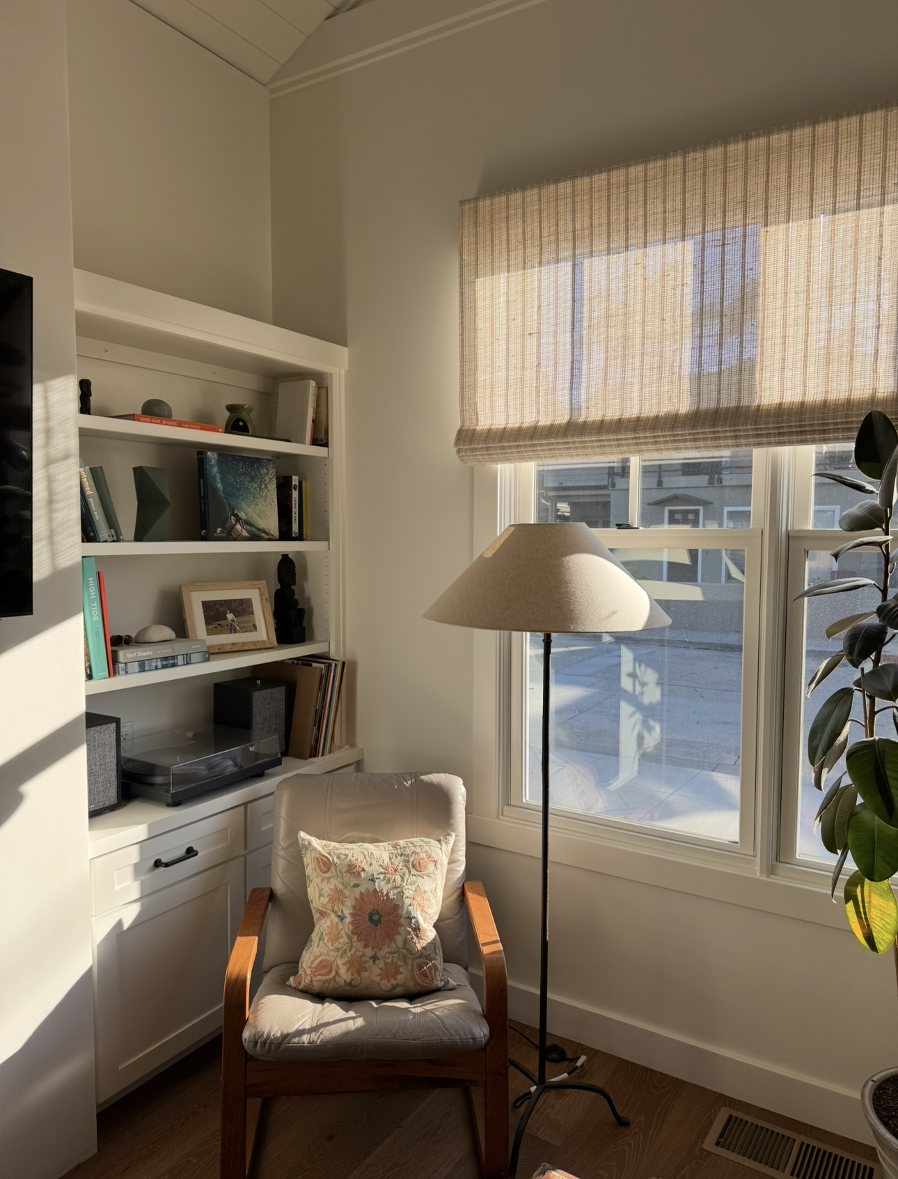 Cozy reading nook with a beige armchair with a floral pillow, a standing lamp, a large window with a beige shade, a white bookshelf filled with books and decorative items, and a potted plant.