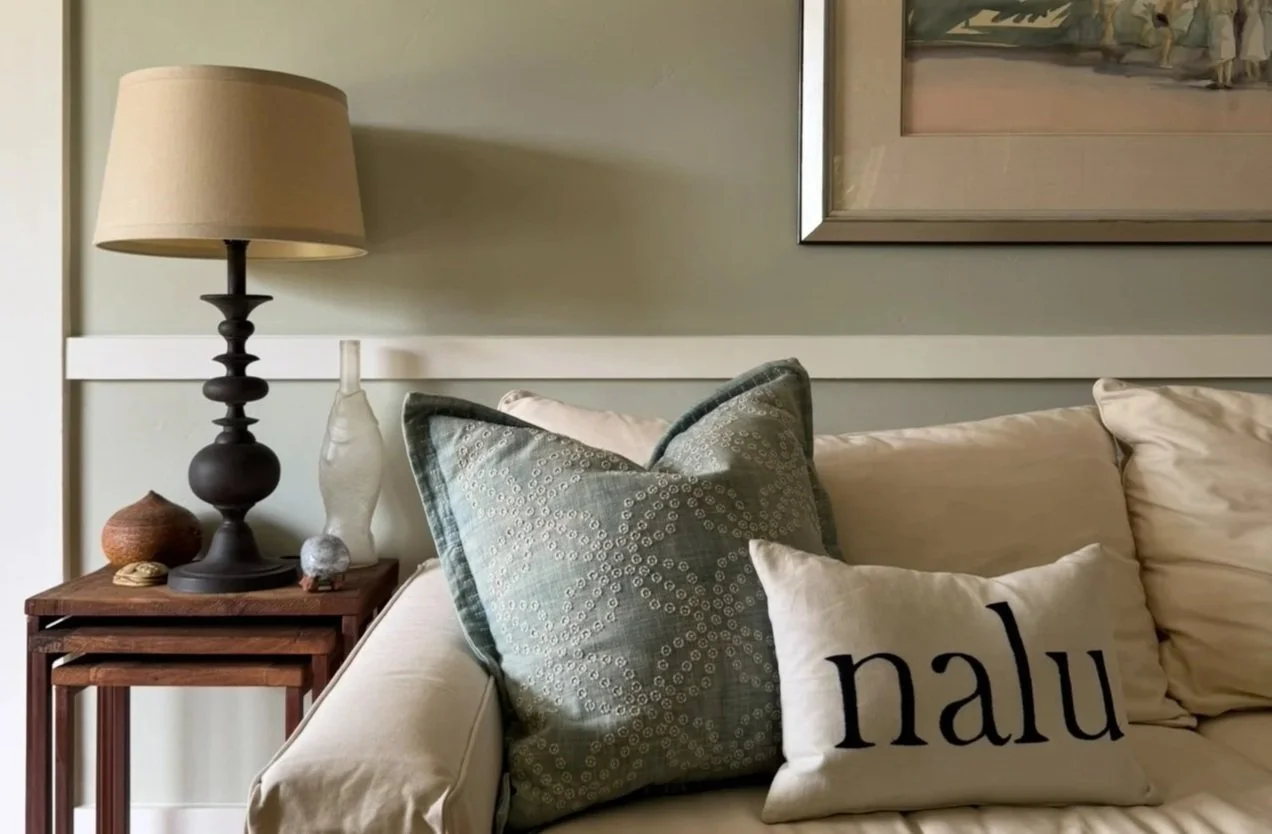 Living room corner with a side table holding a beige lamp, decorative vases, and shells. A couch with patterned and plain pillows, one with the word 'nalu' in black lettering.