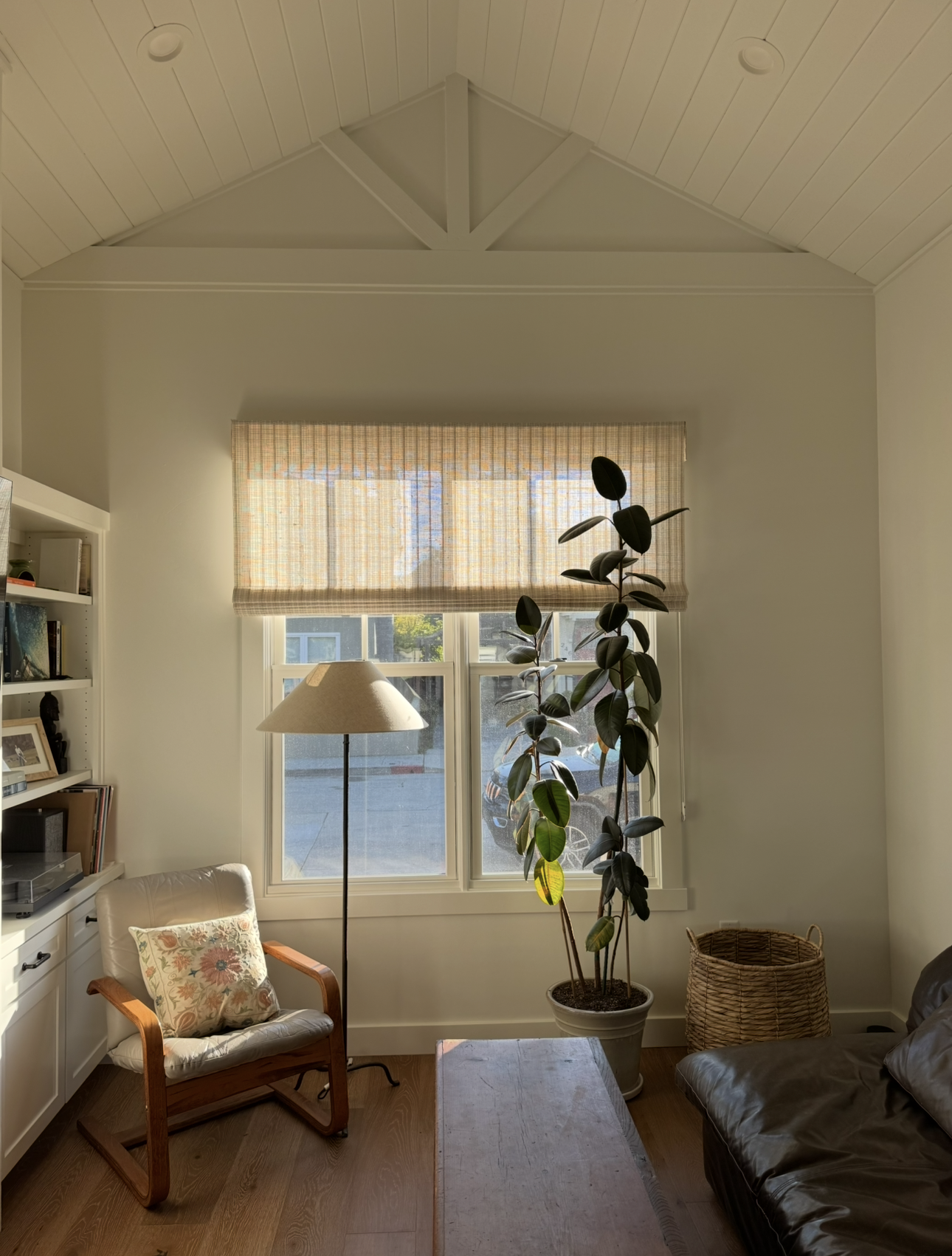 A cozy living room corner with a large window, a floor lamp, a potted plant, a wooden bench, a leather sofa, a cushion, a bookshelf, and a woven basket.