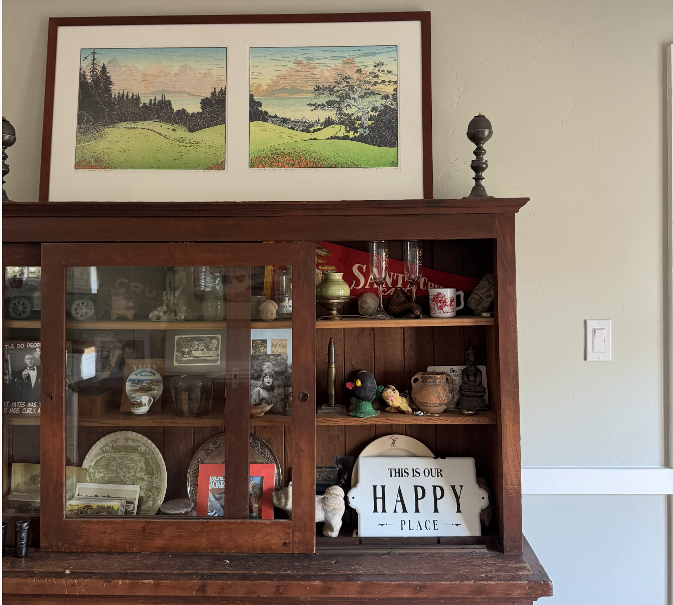 A wooden display cabinet with glass sliding doors filled with various knickknacks, photos, and decorative items. On top of the cabinet, there are two black lamp finials. A framed painting of a landscape with rolling green hills, trees, mountains, and