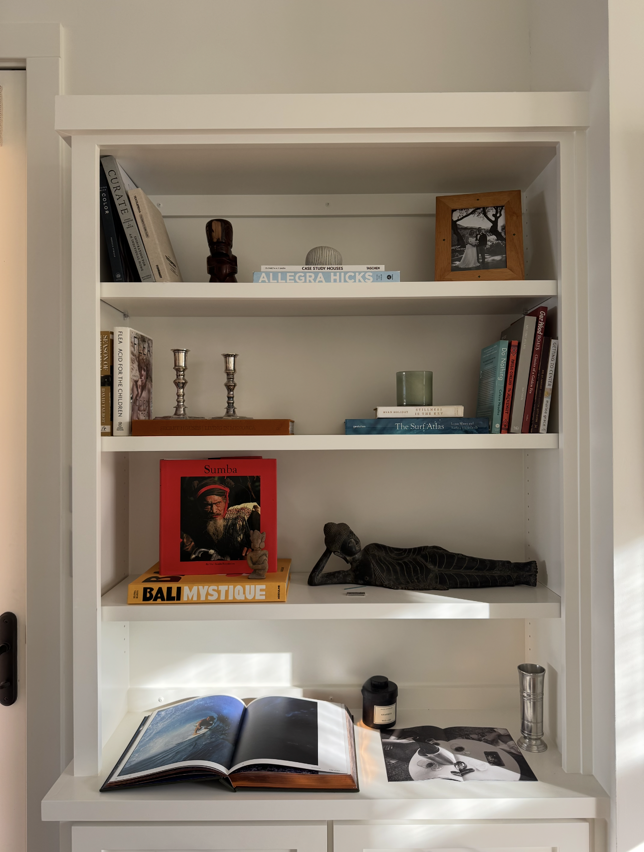 White bookshelf with various books, decorative items, and photographs, including a black and white framed photo, a sculpture, and a magazine on a white surface.
