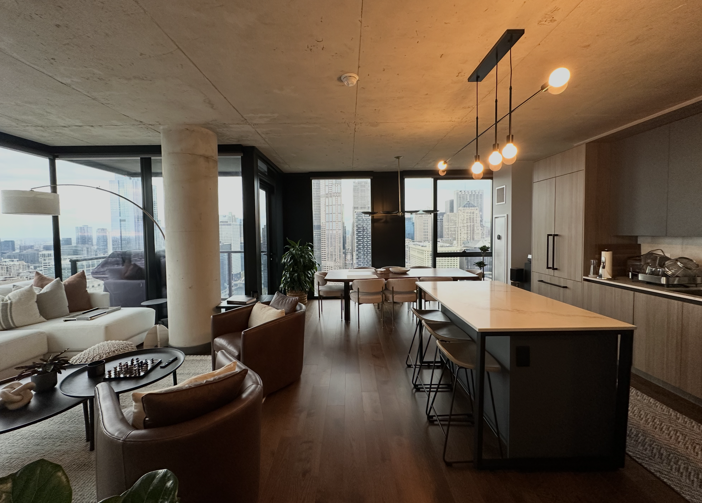 Modern living room and kitchen in a high-rise apartment with city skyline view, featuring a white sofa, leather chairs, a dining table, a kitchen island with stools, and large windows.