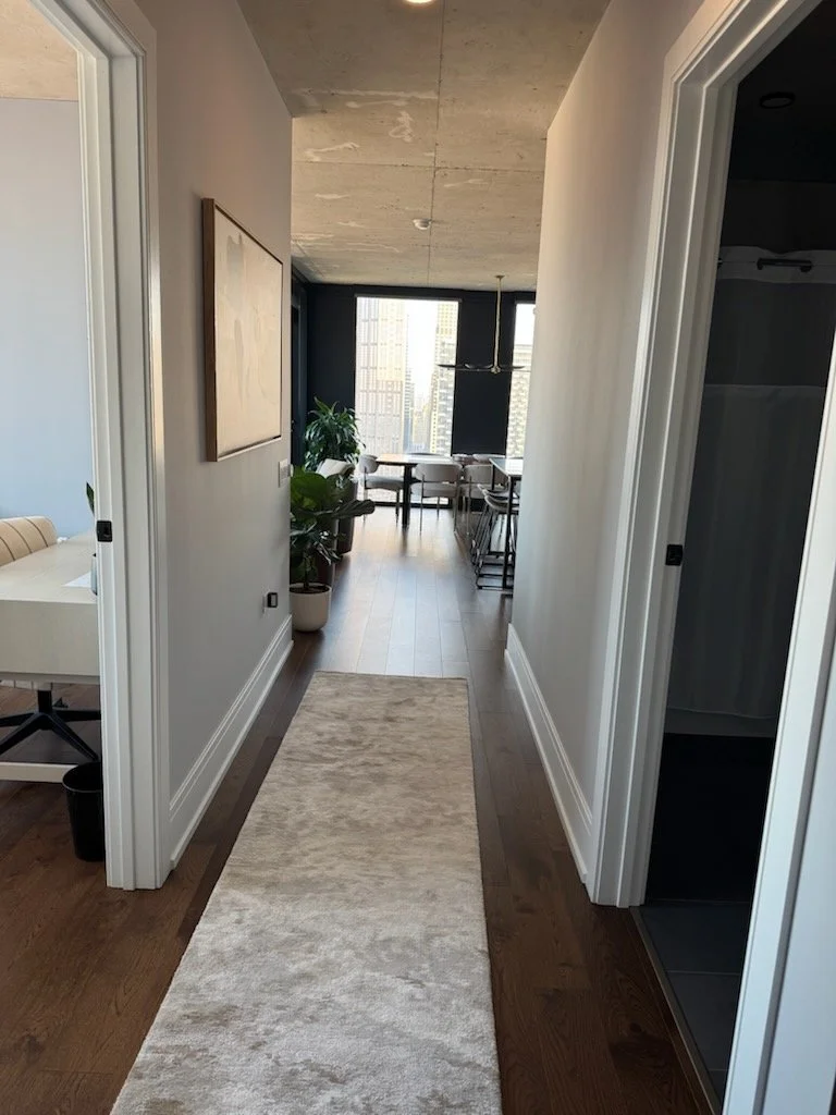 View down a corridor in an apartment with a beige carpet runner, white walls, and a concrete ceiling. At the end, there is a dining area with a table, chairs, and large windows with a cityscape view.