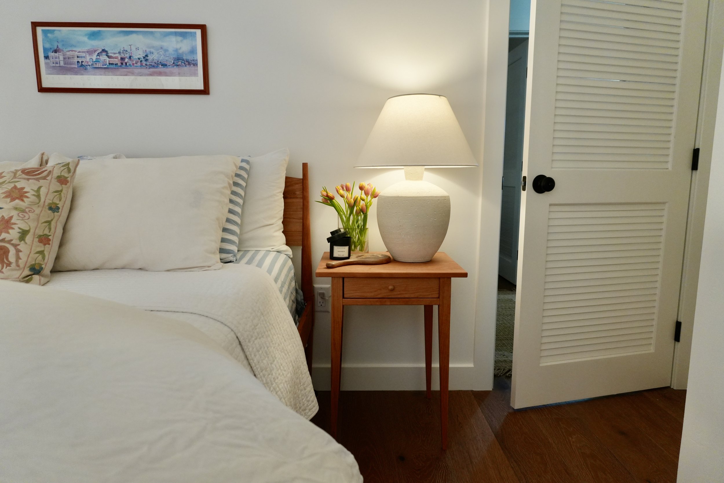 A cozy bedroom corner with a wooden bed, white bedding, decorative pillows, a nightstand with a large ceramic lamp, a small vase of tulips, and a framed artwork on the wall.