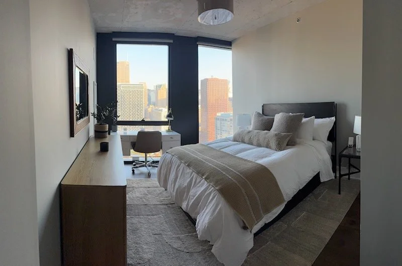 A modern bedroom with a large window view of city buildings, a bed with white bedding and beige accents, a wooden desk with a chair, a wall mirror, and a nightstand with a lamp.