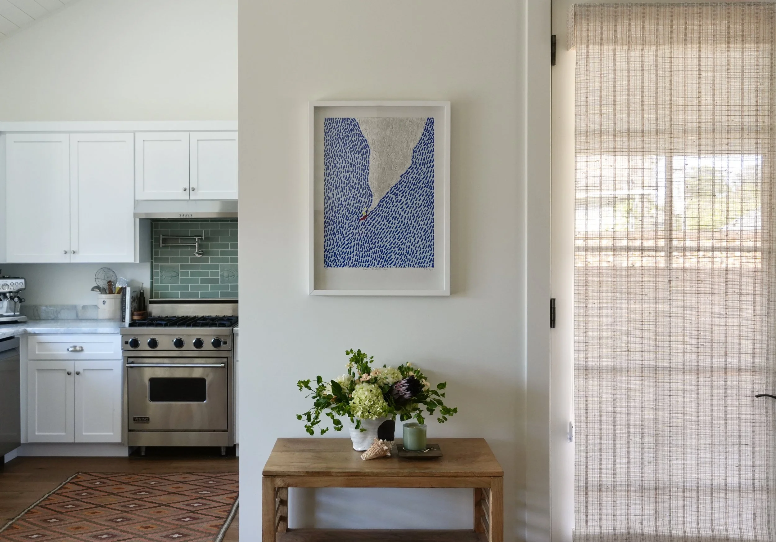 Interior of a kitchen with white cabinets, a stainless steel stove, and a green tiled backsplash. A wooden table with a bouquet of flowers, a candle, and a shell is in front of a white wall with a framed artwork. A door with a woven curtain is on the