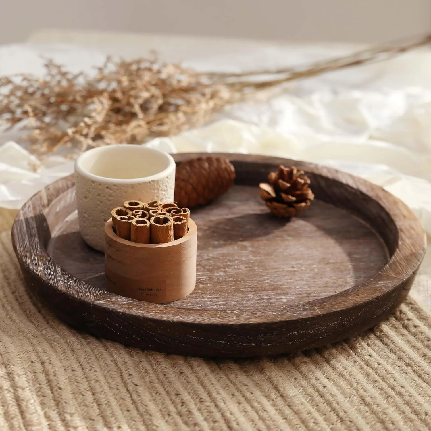 A round wooden tray with a small white textured cup, a bundle of cinnamon sticks, an oak cone, and a pine cone on it, placed on a knitted beige blanket with dried flowers in the background.