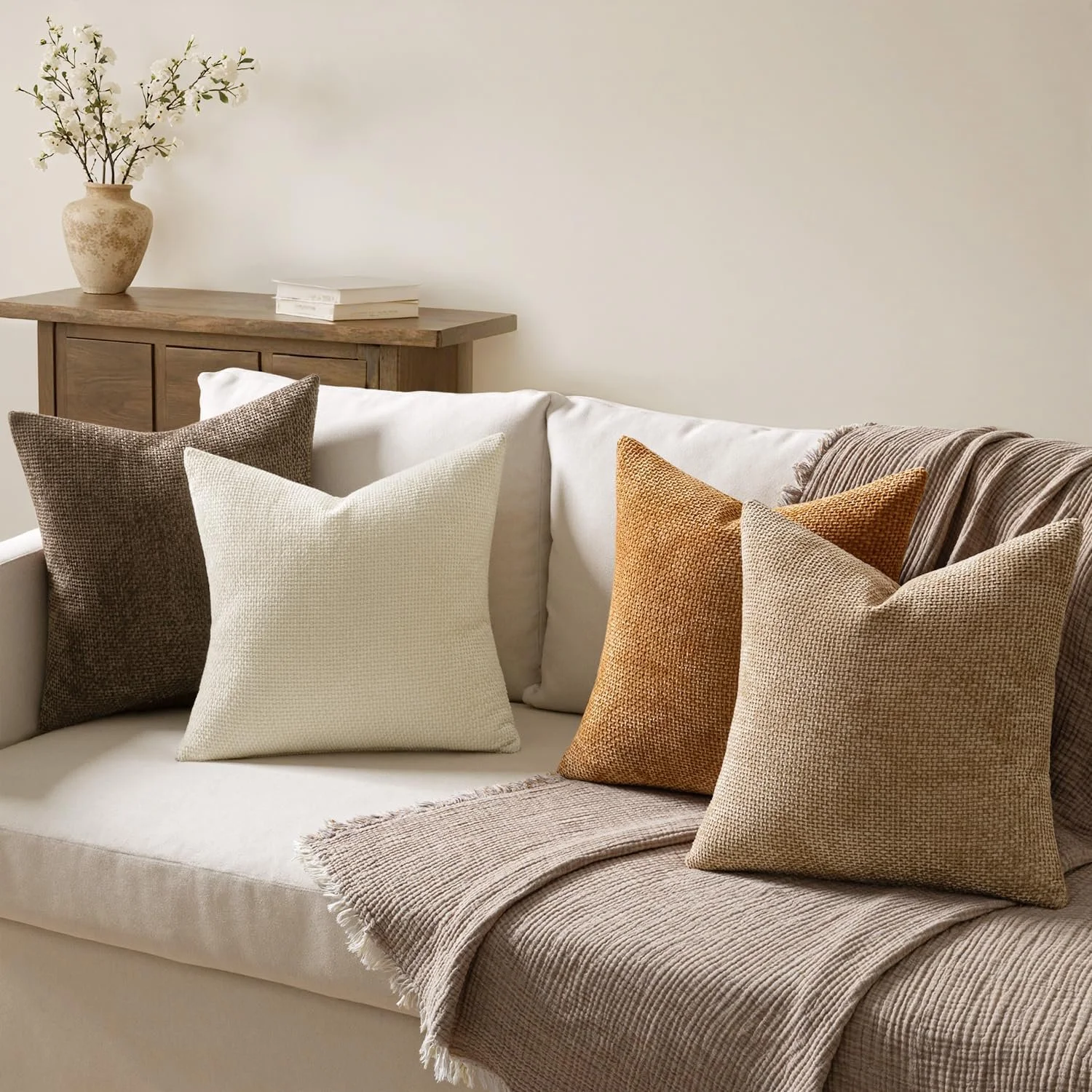 A cozy corner with cushions on a bench and floor, a white textured blanket, a potted plant, and a wooden tray with pinecones and rolled paper.