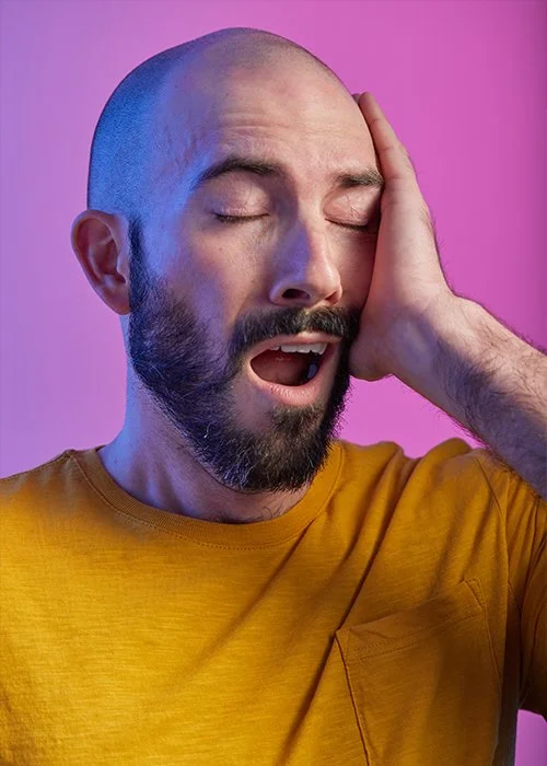 A man with a beard and shaved head yawning or singing with his hand in a phone shape on the side of his face, against a pink and purple background, wearing a yellow shirt.