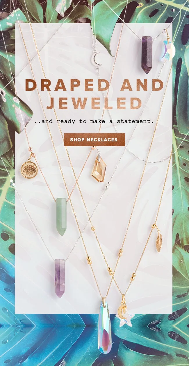 Collection of layered necklaces with pendants, including crystals, moons, and leaves, displayed on a jewelry stand surrounded by green leaves.