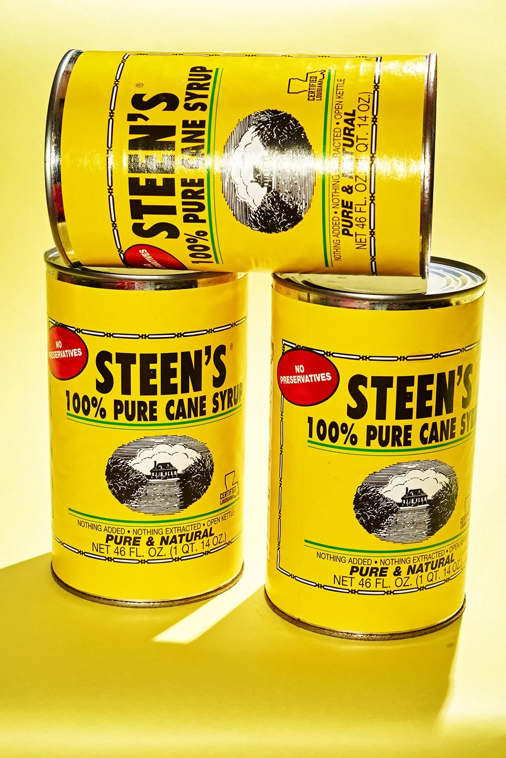 Three cans of Steen's 100% pure cane syrup, yellow labels with black text, one can resting on top of two cans, all cans have metal lids, and a drawing of a house surrounded by trees on the front of each can.