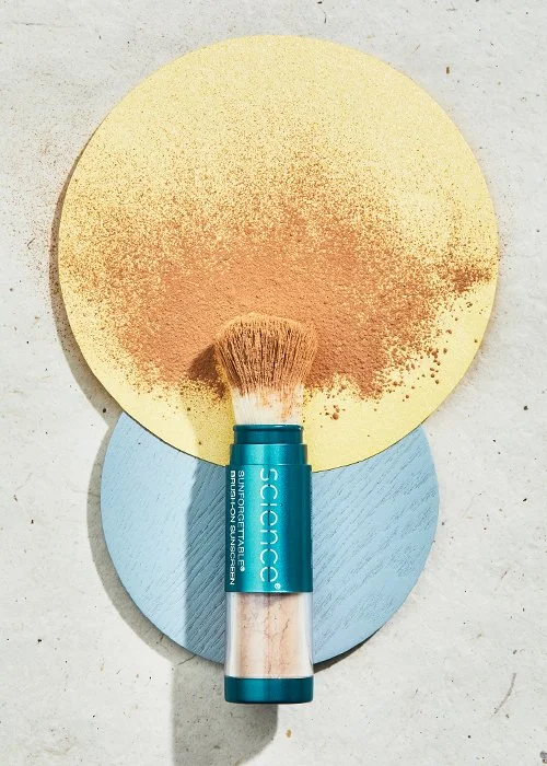 Makeup brush with beige handle and blue ferrule, labeled 'SCIENCE' and 'SILVERTONE BLUSH,' resting on two colored circles, yellow and blue, with powder spilled on the yellow circle.