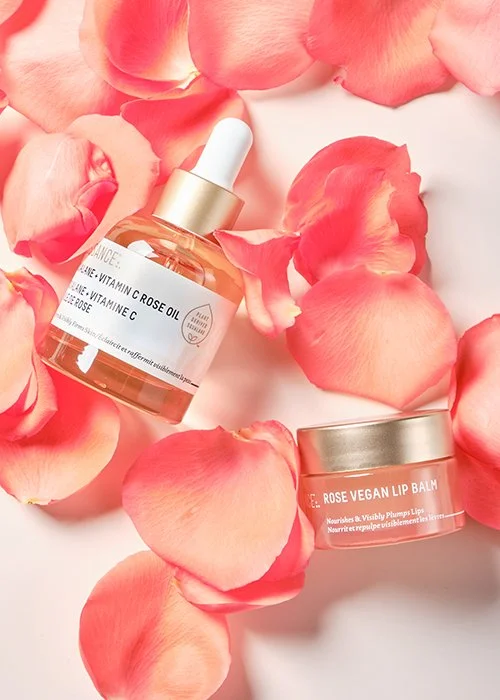 A skincare dropper bottle labeled 'Rose Vitamin C' and a small jar labeled 'Rose Vegan Lip Balm' surrounded by pink rose petals.