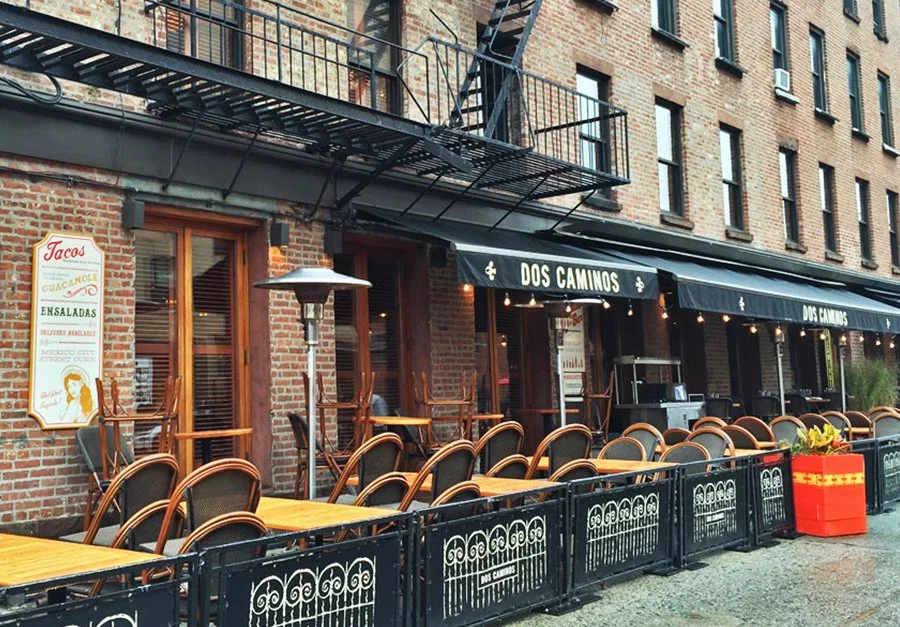 Outdoor seating area of a restaurant called Dos Caminos with empty tables and chairs, a large planter, and a brick building in the background.