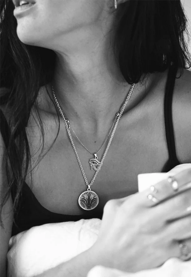 A woman with dark hair wearing layered necklaces, with a close-up of her neck and chest, and holding a phone in her hand. She is dressed in a black strap top.