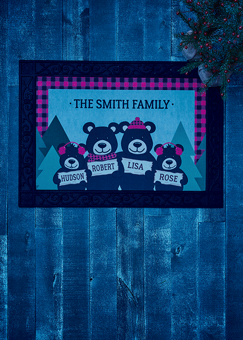 A decorative family-themed wall hanging featuring four bears with winter accessories, each bearing a name: Hudson, Robert, Lisa, and Rose, titled "The Smith Family," hanging on a wooden wall with holiday decorations nearby.