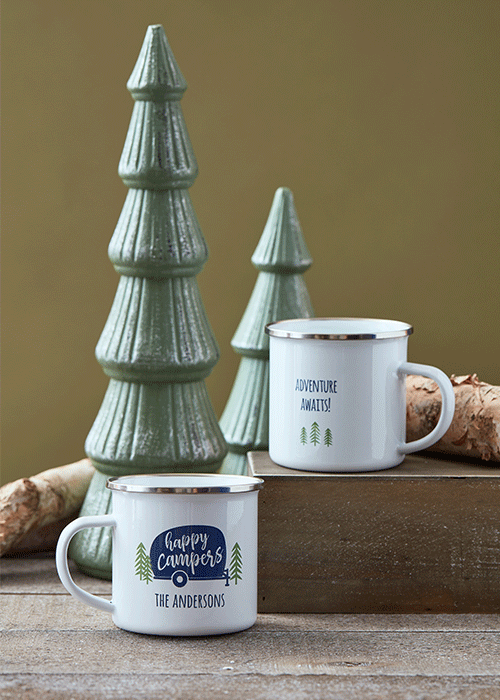Two white camping mugs with green and blue designs, one saying "Happy Campers The Andersons" and the other saying "Adventure Awaits!", placed on a wooden surface with ceramic trees in the background.