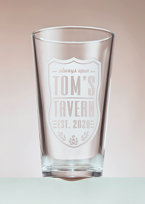 A clear glass with a white logo that reads 'always open TOMS TAVERN EST. 2020' on a light background.