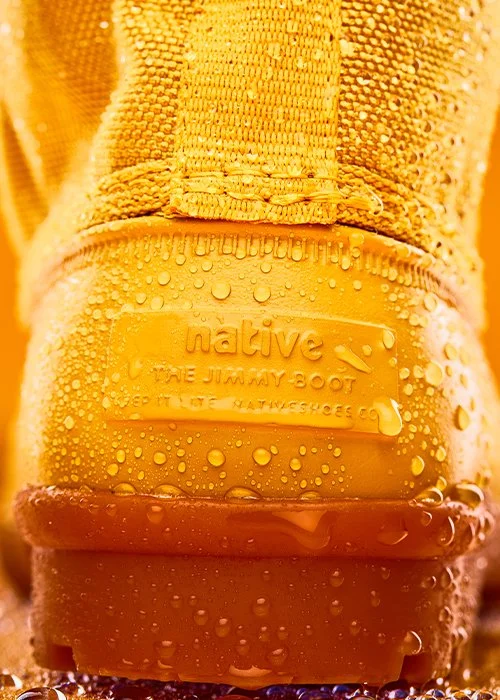 Close-up of a yellow Native brand hiking boot with water droplets on it, displaying the brand logo and size, set against a colorful background.