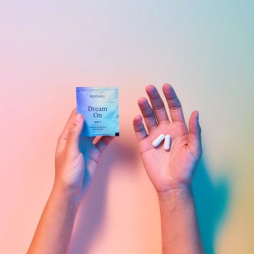 A person holding a blue box labeled "Dream On" and three white pills on their other hand against a pastel pink and blue background.