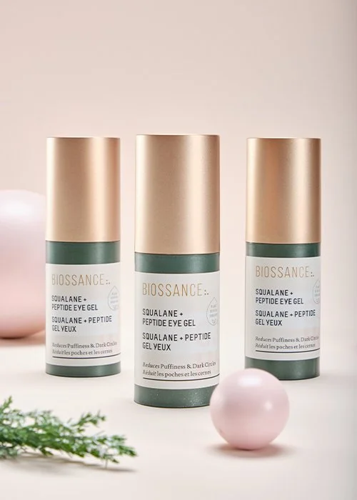 Three bottles of Biossance Squalane + Peptide Eye Gel, with a pink sphere and some green foliage in the foreground on a white surface against a light background.