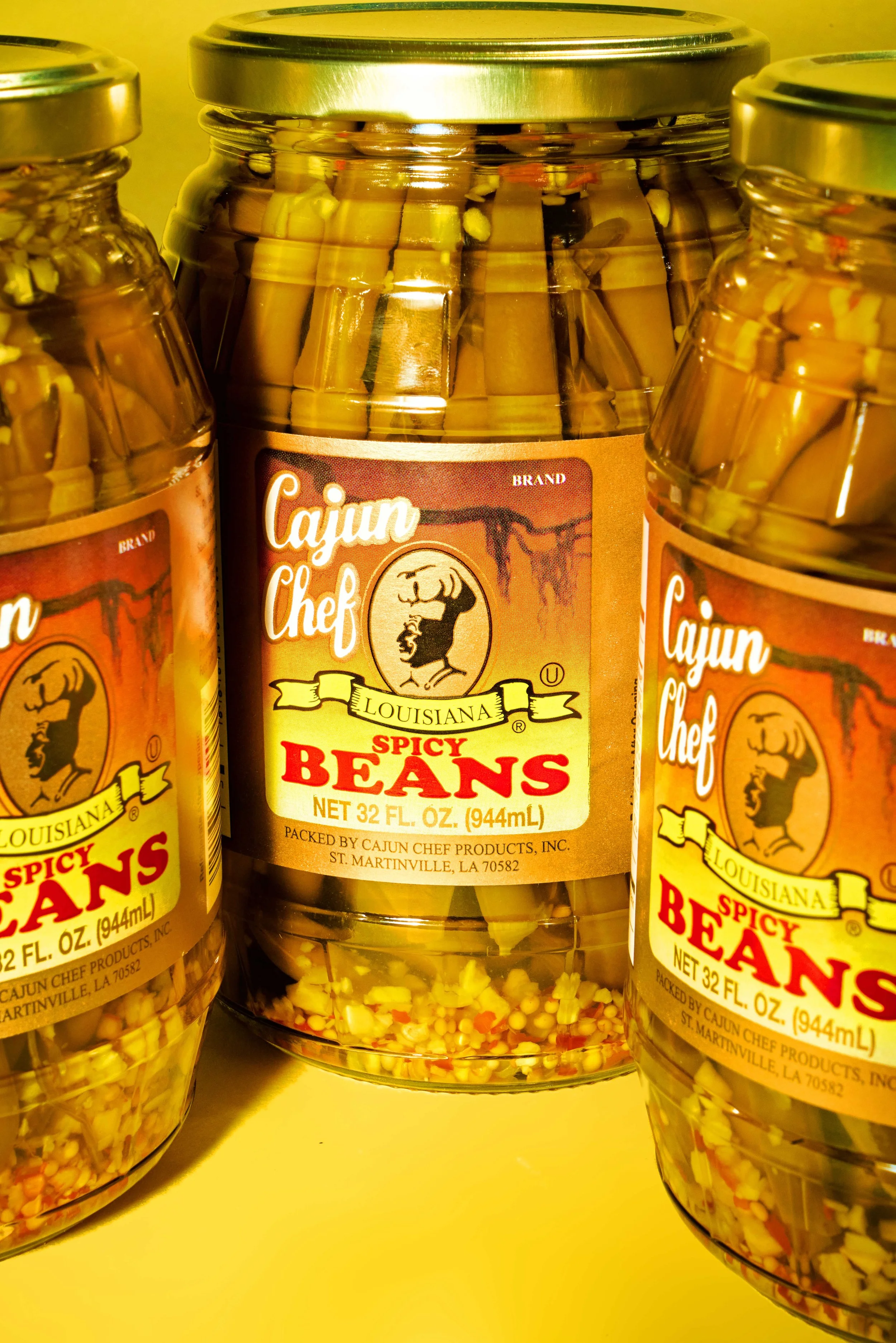 Three jars of Cajun Chef Louisiana spicy beans with yellow and red seasoning inside, placed on a yellow surface.
